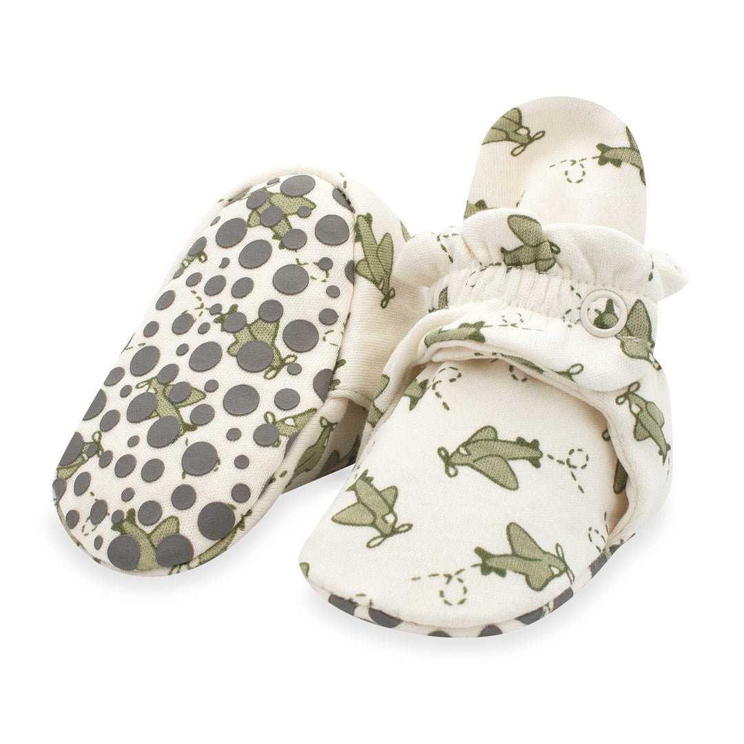 A pair of organic cotton baby booties with gripper soles, displayed against a white background. The booties have a green airplane print on a cream base.