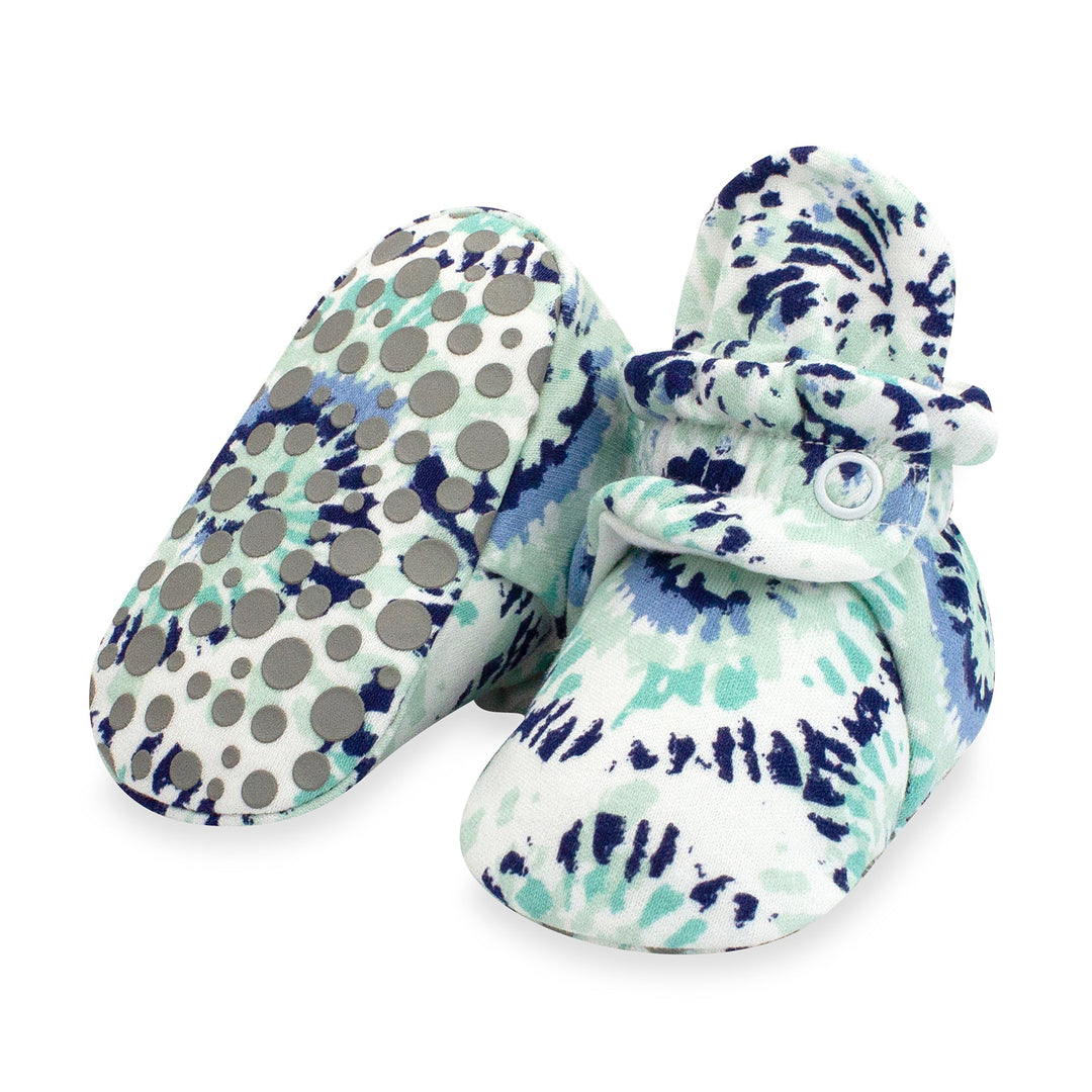 A pair of organic cotton baby booties with a tie-dye pattern in shades of blue and white, featuring rubber grippers on the bottom.