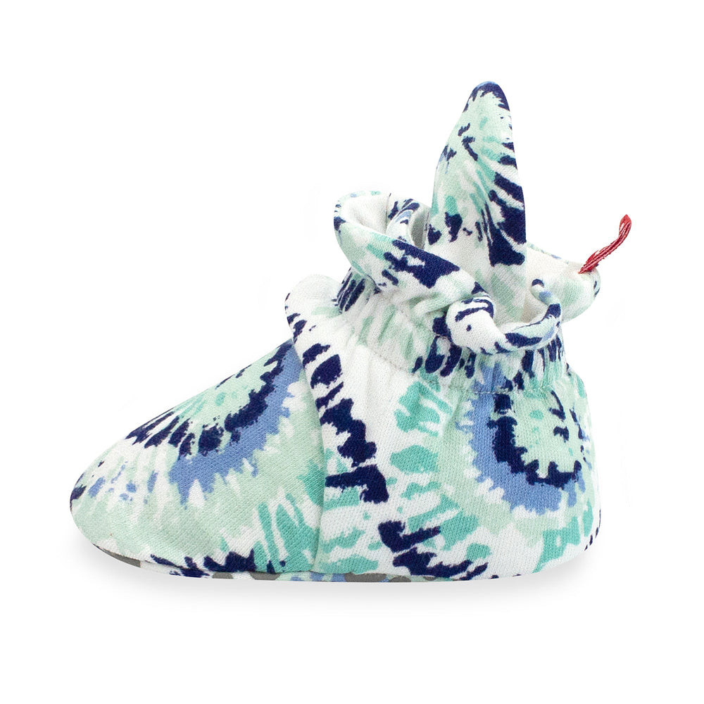 A pair of organic cotton baby booties with a tie-dye pattern in shades of blue and white, featuring rubber grippers on the bottom.