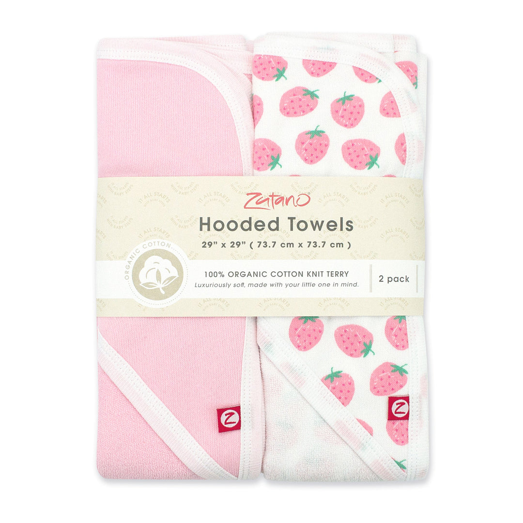 Two packaged hooded towels for babies, one with a strawberries print and the other in baby pink with white trim, both on a pink background.