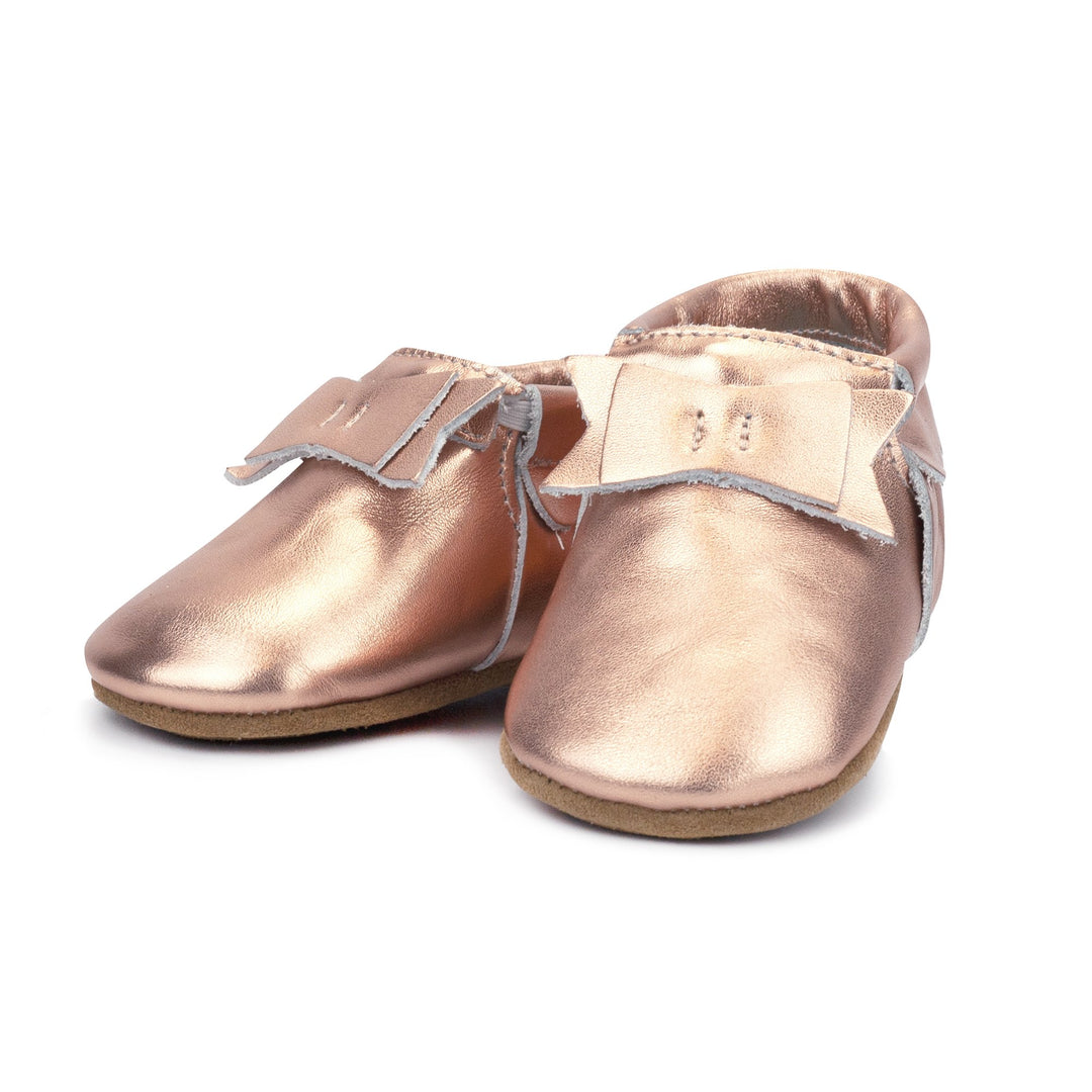 A pair of metallic rose gold leather baby moccasins with a bow on the tongue and a non-slip sole.