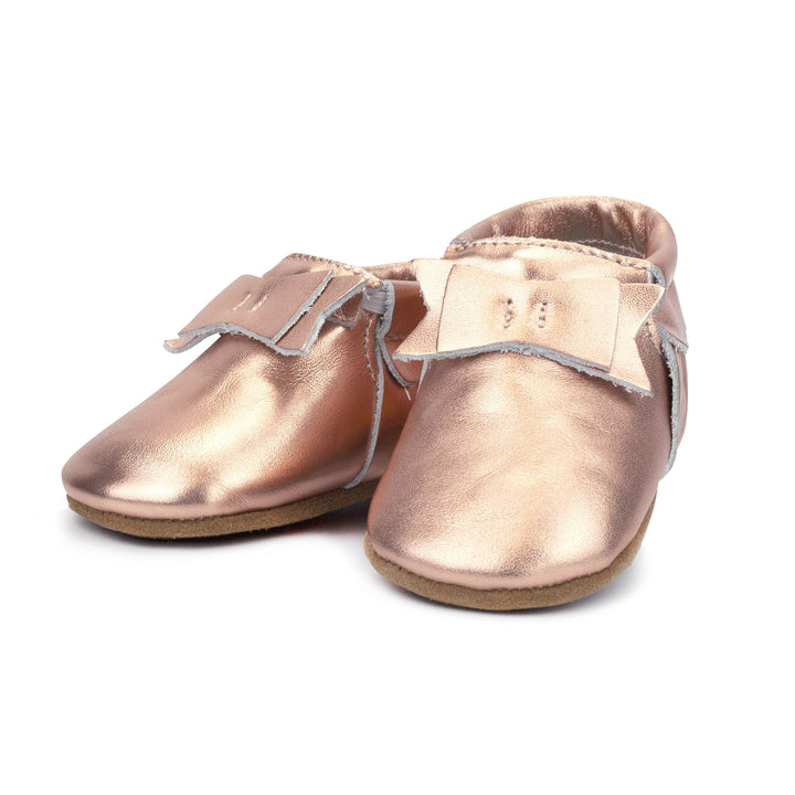 A pair of metallic rose gold leather baby moccasins with a bow on the tongue and a non-slip sole.