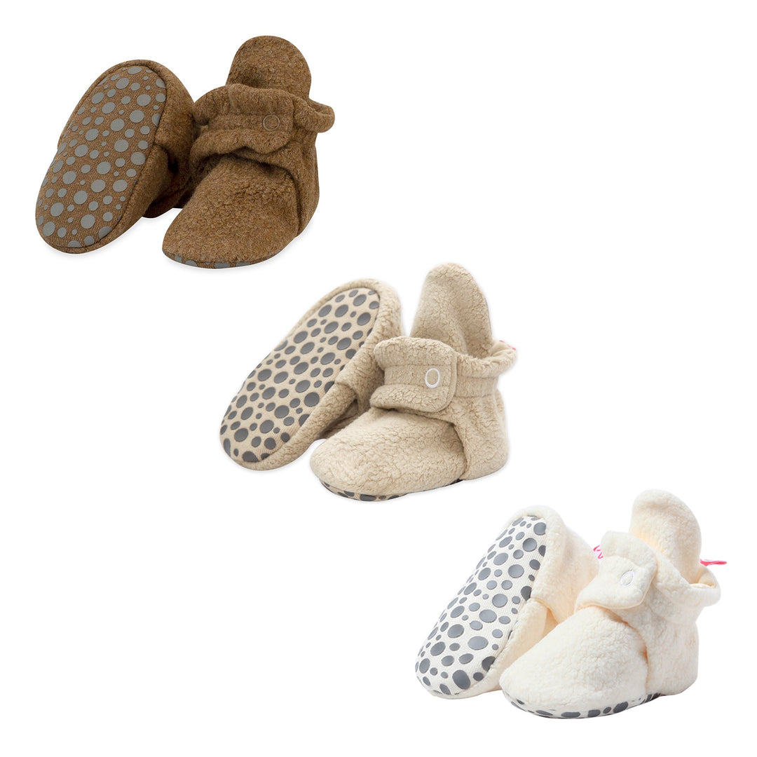 Three pairs of Cozie fleece gripper baby booties in a three pack, featuring heather mocha, khaki, and cream colors. The booties have adjustable snaps and gripper bottoms.