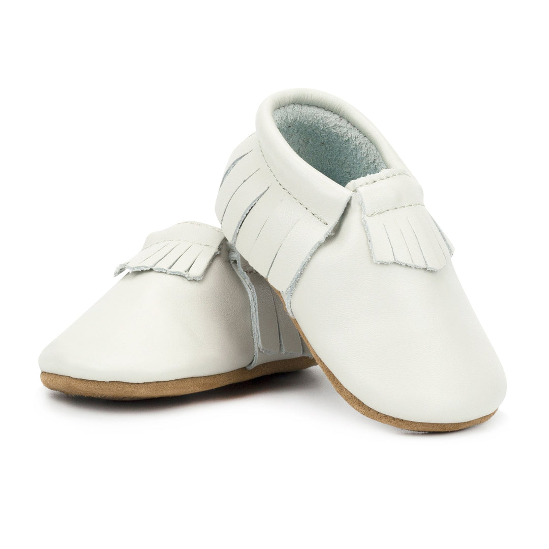 A pair of white leather baby moccasins with fringe detailing on the tongue, sides, and heel.