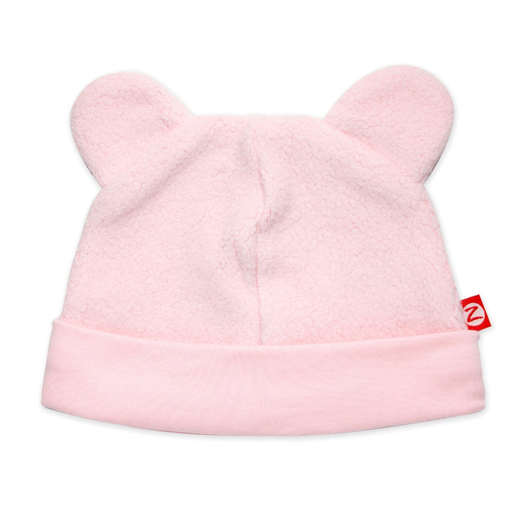 A baby pink fleece winter hat with ears designed to resemble a bear's face.