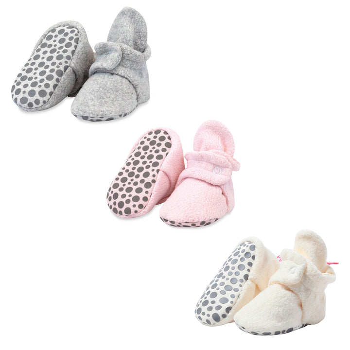 Three pairs of baby booties in a three pack, featuring heather gray, baby pink, and cream colors with gripper bottoms.