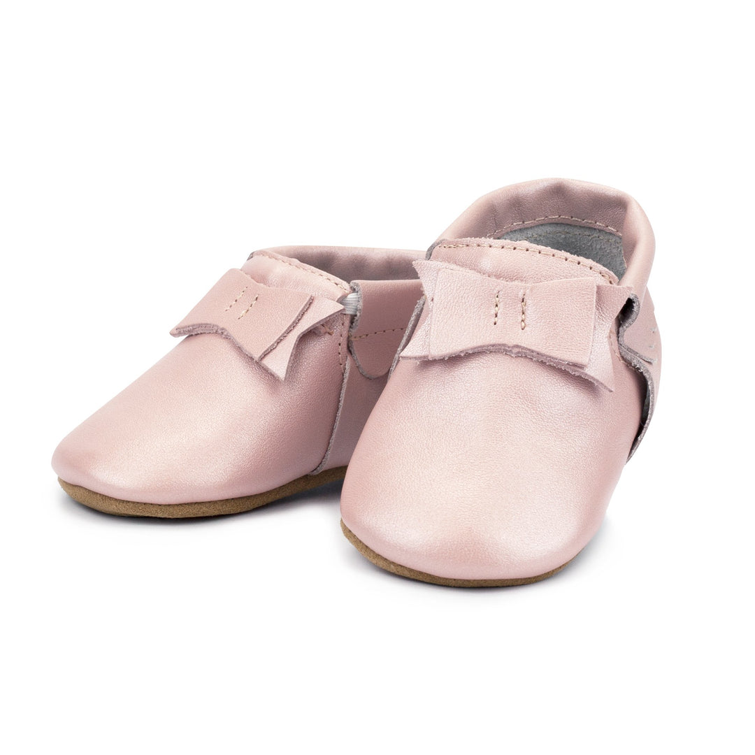 A pair of pink leather baby moccasins with a bow on the tongue and a non-slip sole.